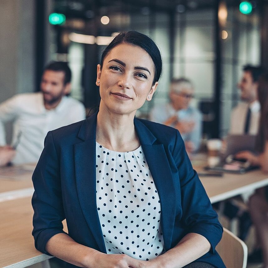 Selbstbewusste Geschäftsfrau in elegantem Business-Outfit sitzt in moderner Meeting-Umgebung und blickt vorausschauend