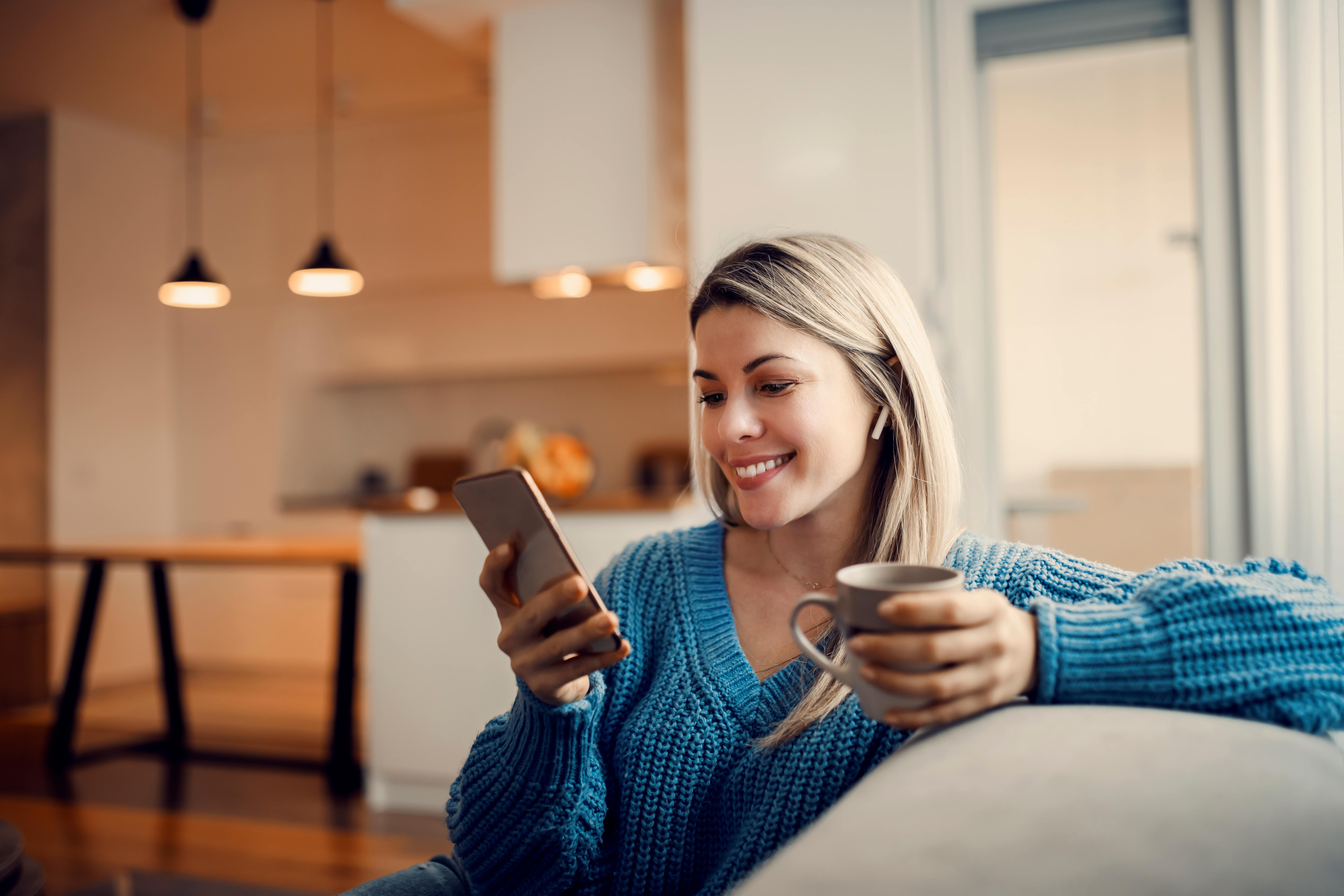 Lächelnde Frau in gemütlicher Umgebung mit Smartphone und Kaffeetasse