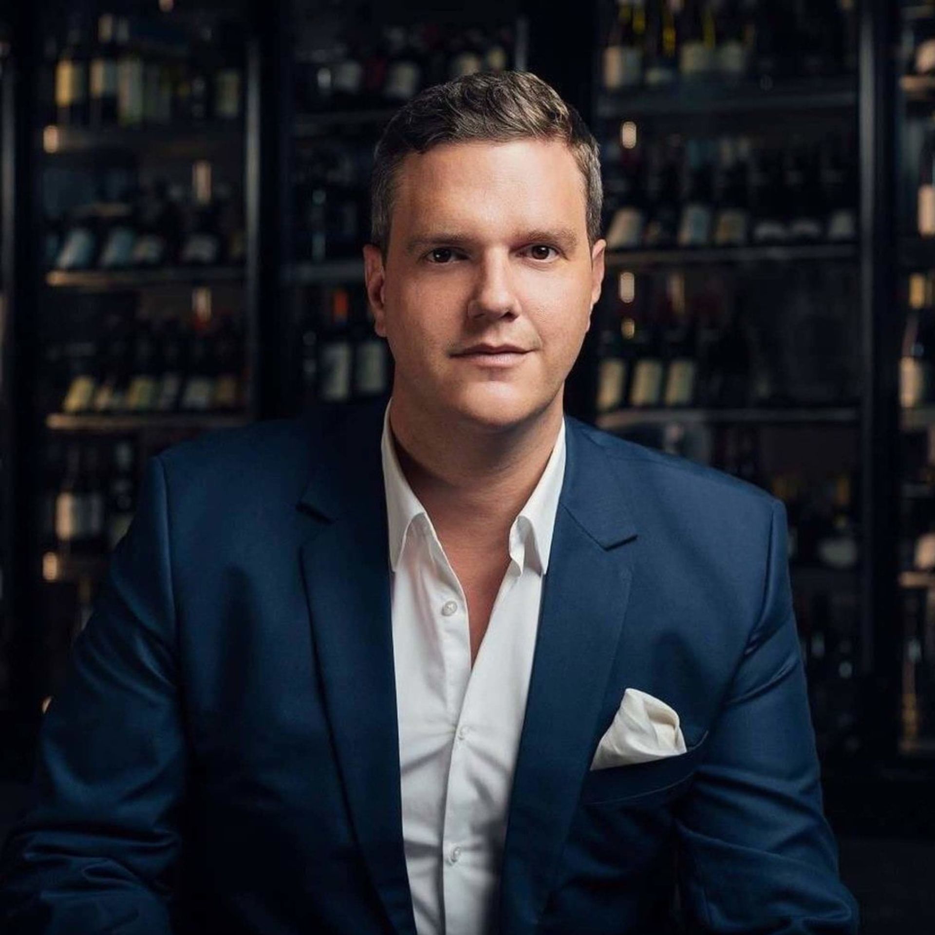 Oliver Schreiber, Eigentümer des Best Western Plus Atrium Hotel in Ulm, in elegantem dunkelblauen Anzug vor einer stilvollen Weinregal-Kulisse. Sein selbstbewusstes Auftreten unterstreicht seine Leidenschaft für exzellenten Service und gehobene Gastlichkeit