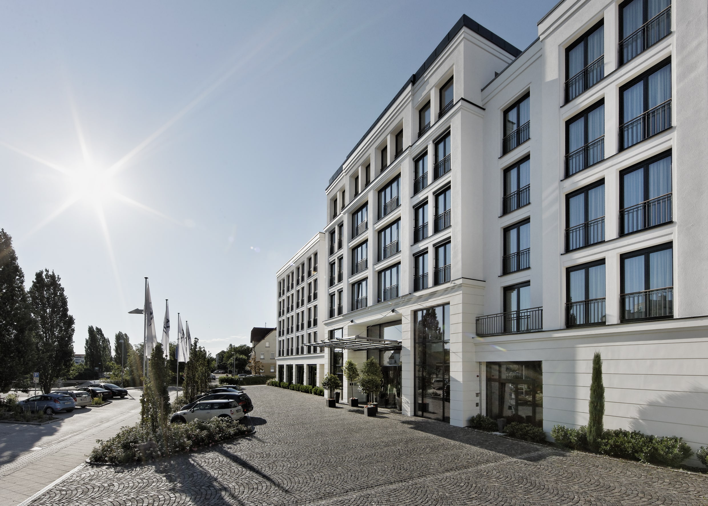 Elegante Hotel-Fassade in zeitloser Architektur mit großen Fenstern und heller Steinverkleidung. Die weitläufige, gepflasterte Eingangszone mit gepflegten Pflanzen verleiht dem Gebäude ein einladendes Ambiente. Strahlender Sonnenschein unterstreicht die moderne und freundliche Atmosphäre des Hotels